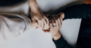This article is about preventing suicidal thoughts. Two people hold each others hands on top of a table, one is wearing a white jumper and the other is wearing a black jumper