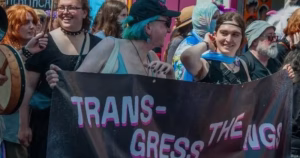 The article is about Transgress the NGS open letter. The image depicts a crowd protest and two people holding a banner that says Transgress the NGS
