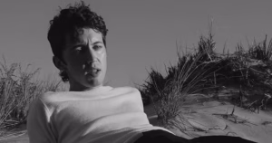 Troye Sivan in the music video for FF. It is a black and white image of Sivan lying on a beach amongst the dunes.